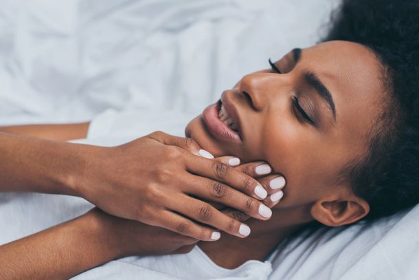 woman with jaw popping and stiffness, holding her jaw, wincing in pain, woman laying in bed