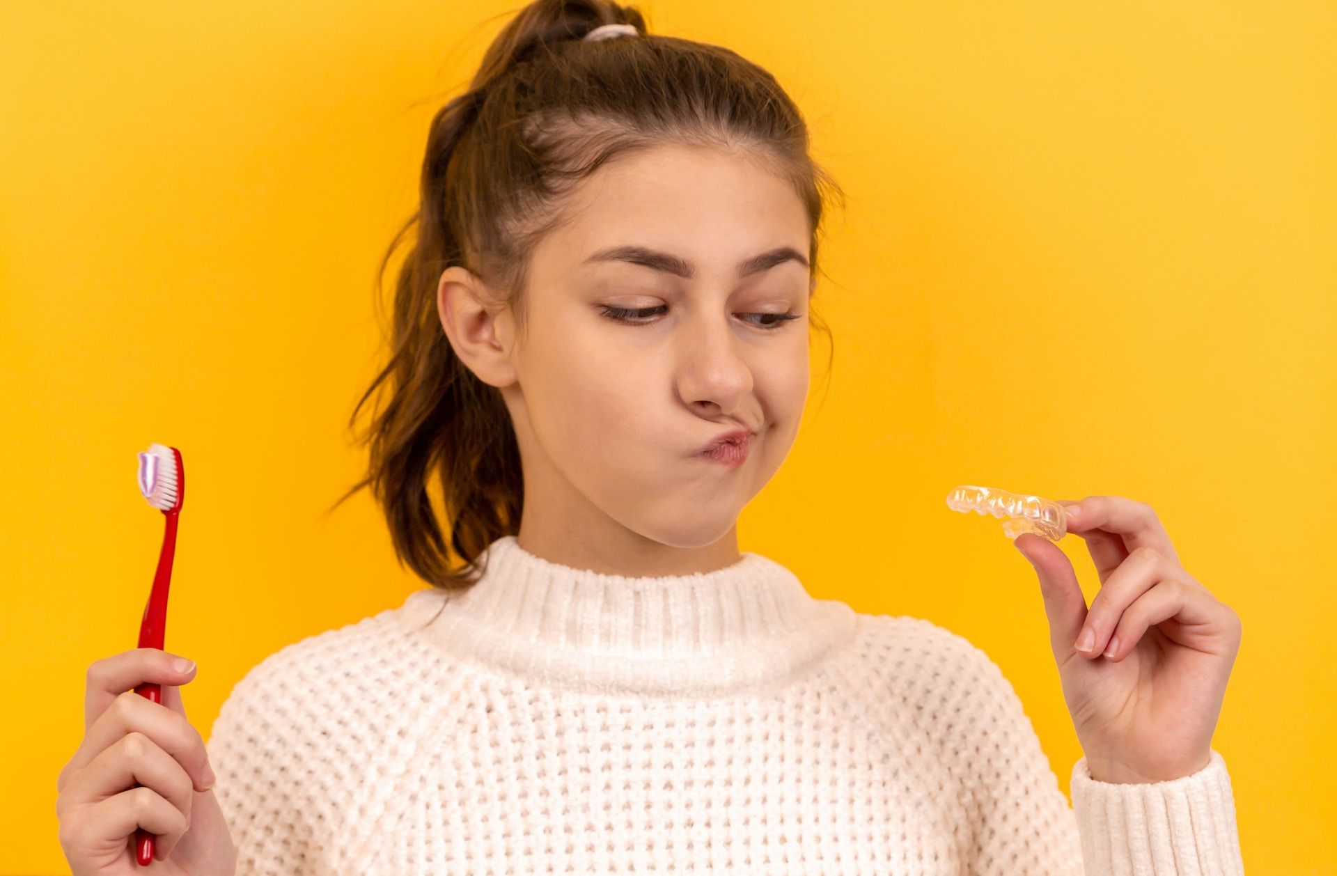 woman holding clear aligner or retainer and toothbrush on a yellow background